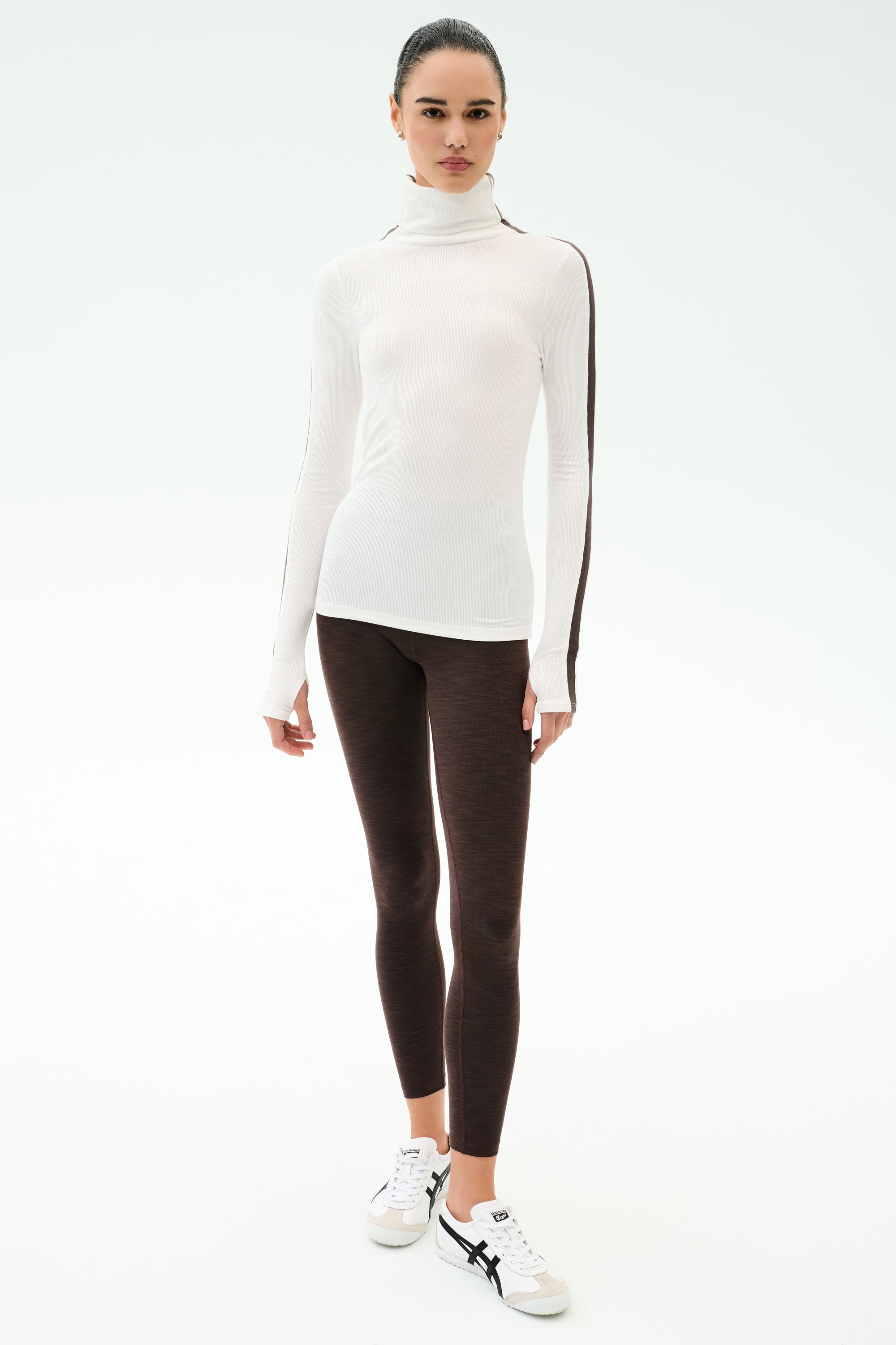 A woman faces forward in the SPLITS59 Jackson Rib Full Length Turtleneck in White/Dark Chocolate, paired with dark leggings and white sneakers—perfect for yoga or running, set against a plain white background.