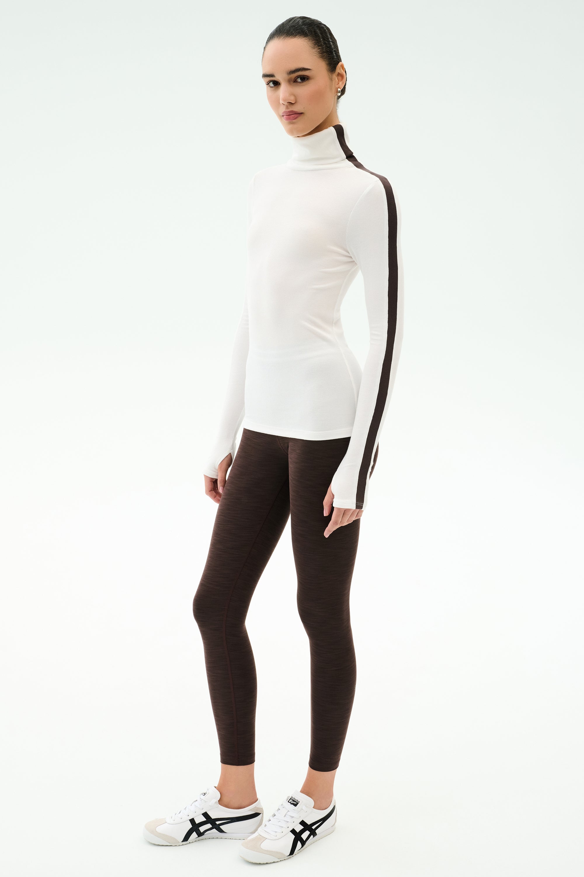 A woman stands against a plain background wearing the SPLITS59 Jackson Rib Full Length Turtleneck in White/Dark Chocolate, brown leggings, and white athletic shoes—ideal for stylish yoga or running sessions.