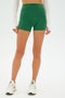A person in the SPLITS59 Ella Airweight High Waist Short 3.5" in Arugula/White, with a white long-sleeve crop top and sleek white sneakers, stands elegantly against a plain background.