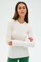 A serene yoga-inspired person with long brown hair stands against a plain backdrop, wearing the SPLITS59 Louise Rib Long Sleeve in White and green pants, arms crossed.