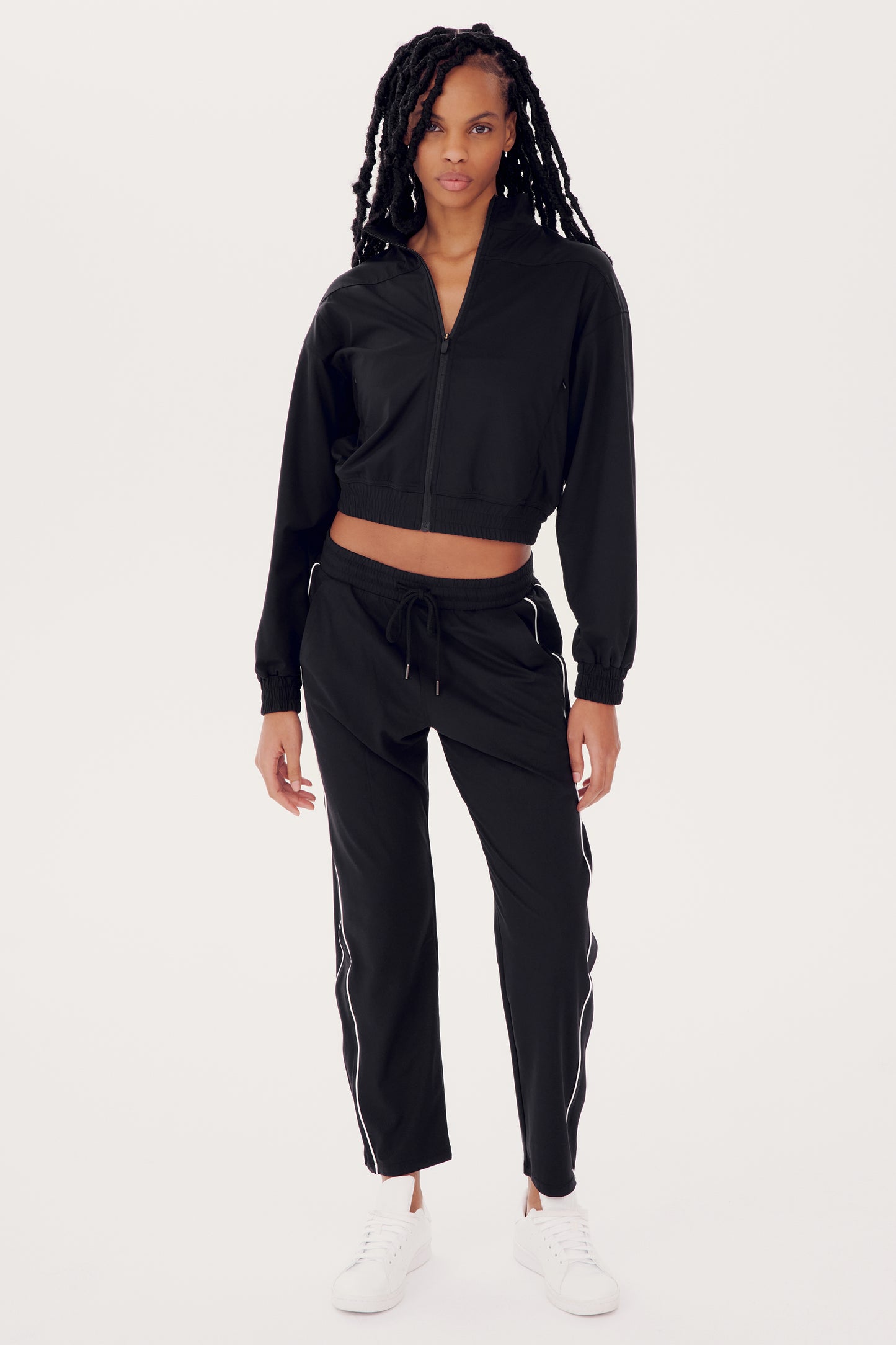 A person is dressed in the SPLITS59 Lucy Rigor Pant W/Piping in black with white stripes and white sneakers, standing on a plain white background. The relaxed-fit jogger ensures all-day comfort, ideal for any activity.