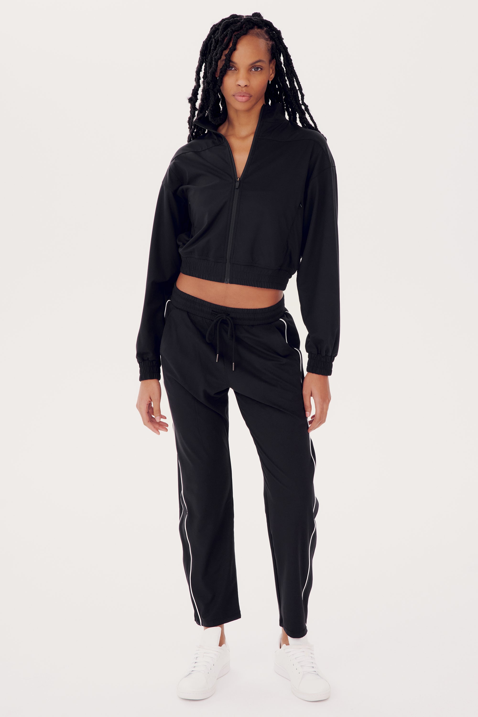 A person is dressed in the SPLITS59 Lucy Rigor Pant W/Piping in black with white stripes and white sneakers, standing on a plain white background. The relaxed-fit jogger ensures all-day comfort, ideal for any activity.