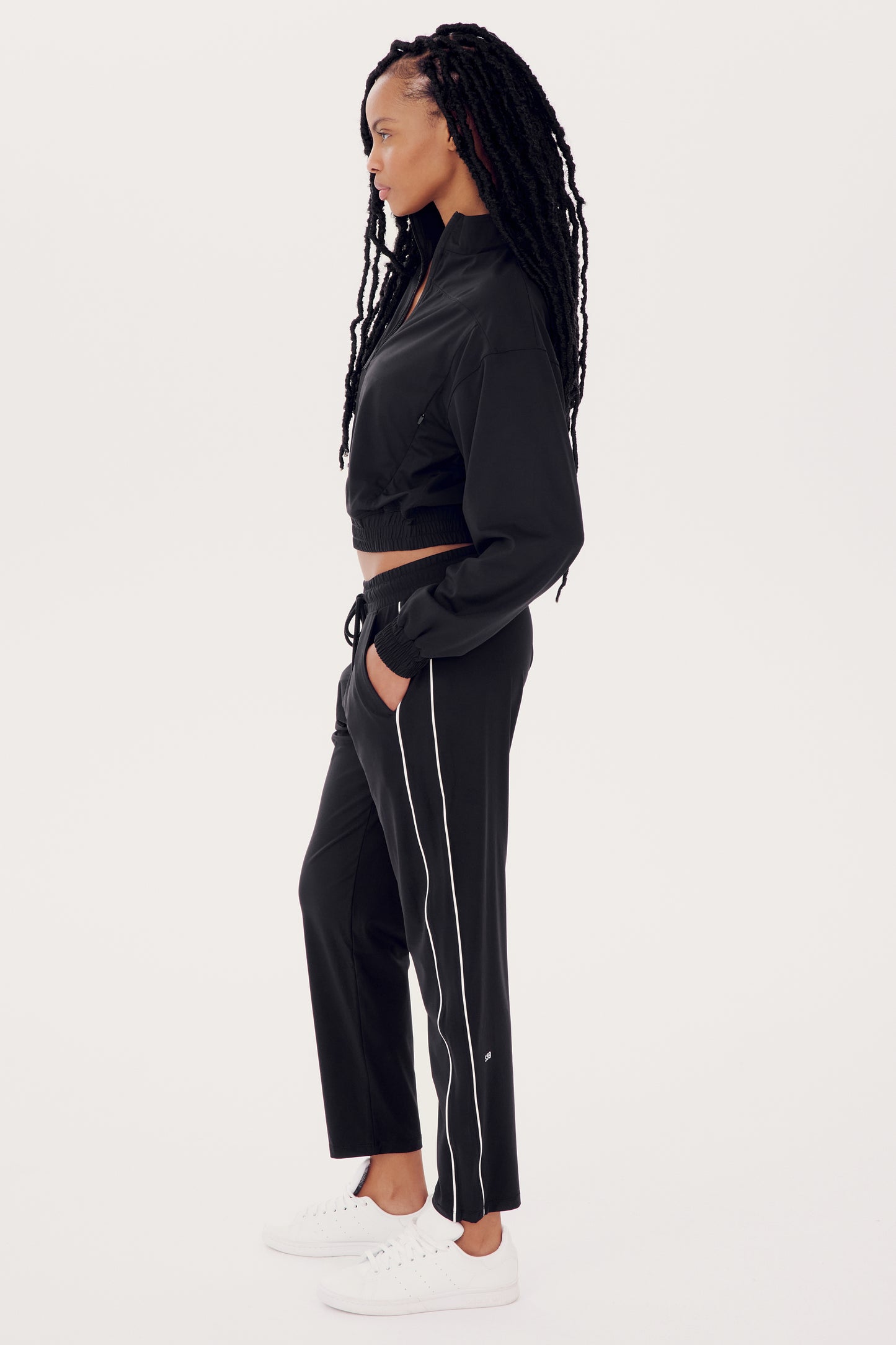 Wearing the SPLITS59 Lucy Rigor Pant W/Piping in black with white side stripes and white sneakers, a person in profile against a plain background exudes all-day comfort with this relaxed fit jogger design that ensures ease of movement and style.