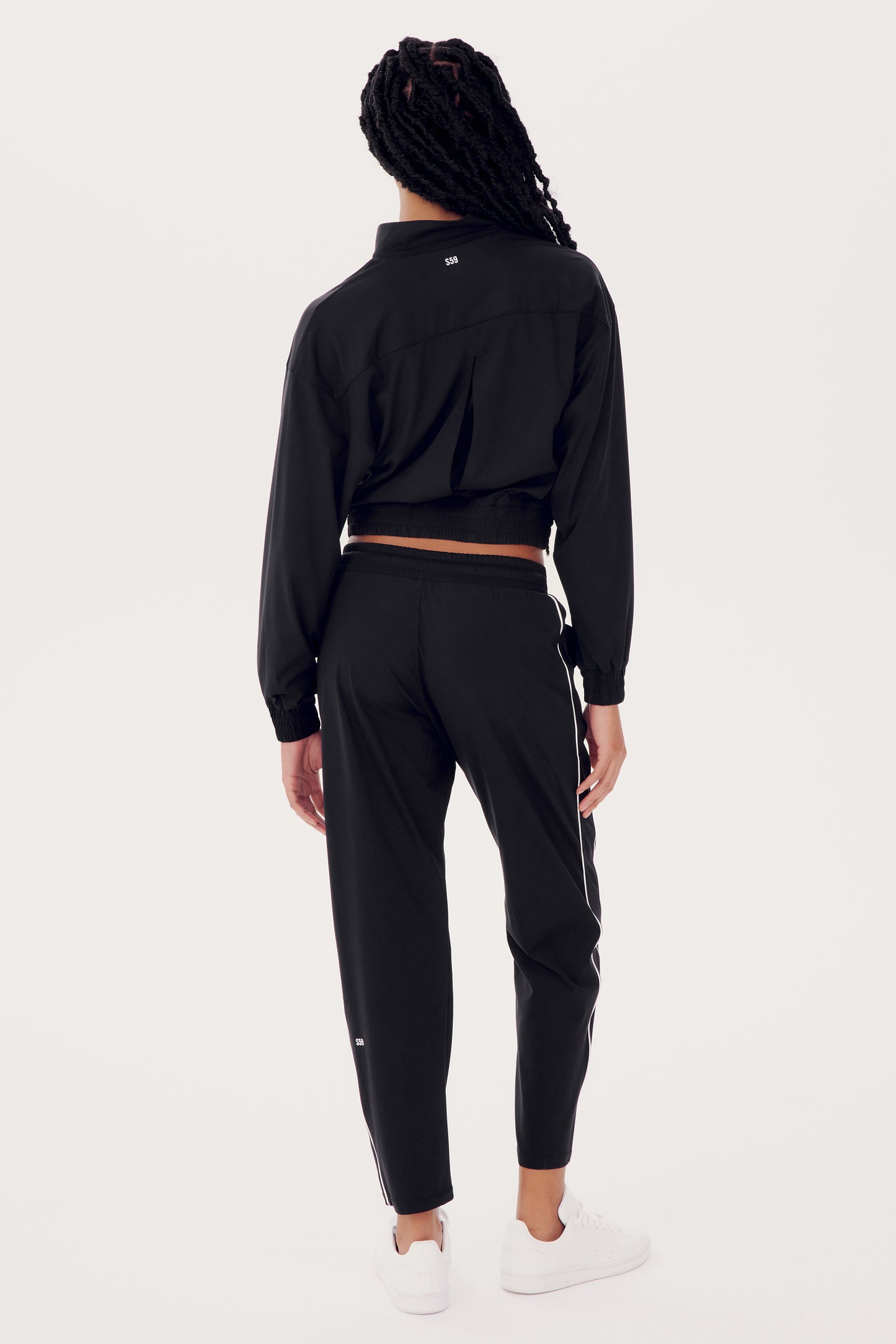 Someone wearing the SPLITS59 Lucy Rigor Pant W/Piping in black and white sneakers stands against a plain background, with their back to the camera, exuding all-day comfort.