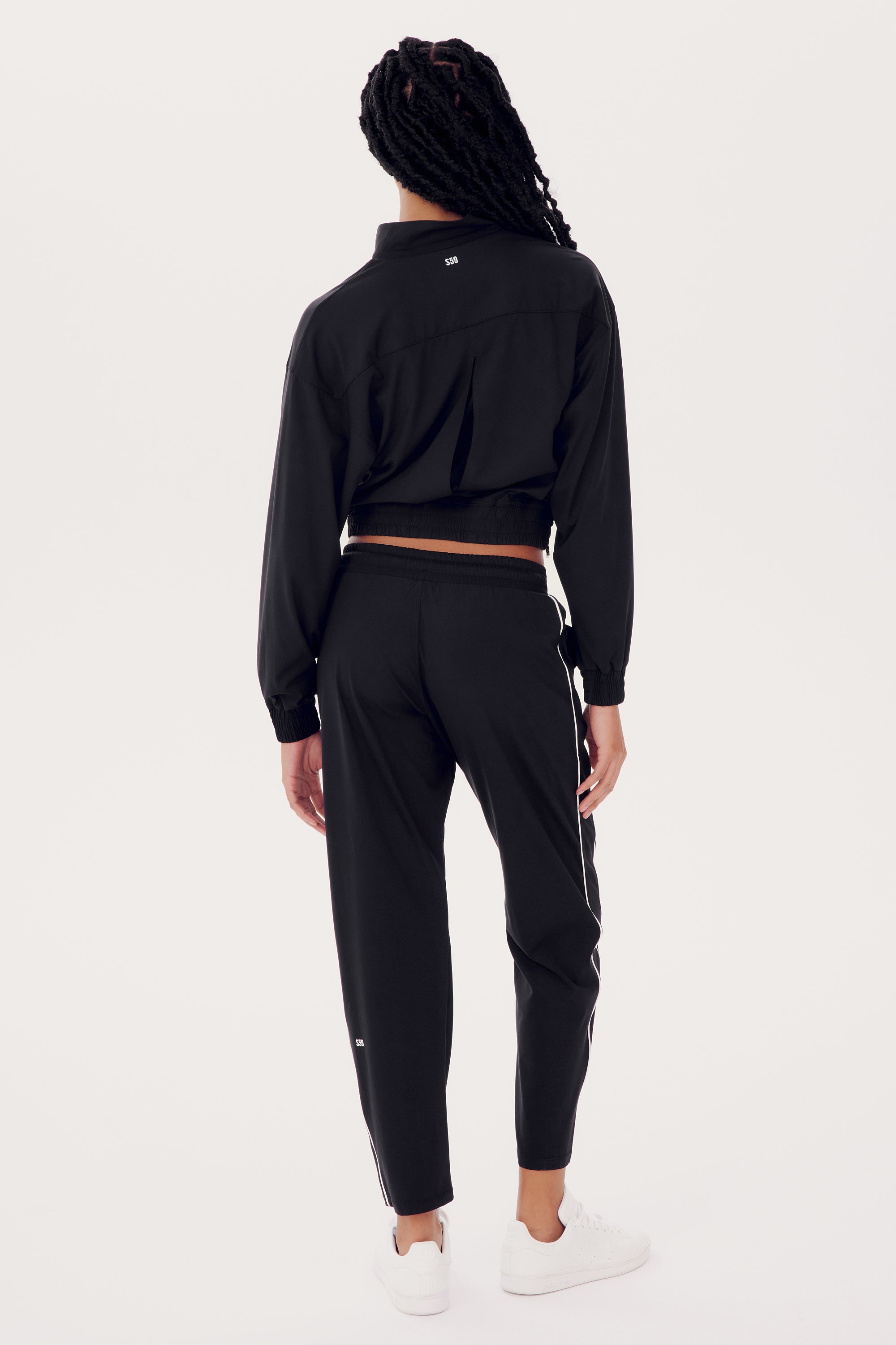 Someone wearing the SPLITS59 Lucy Rigor Pant W/Piping in black and white sneakers stands against a plain background, with their back to the camera, exuding all-day comfort.