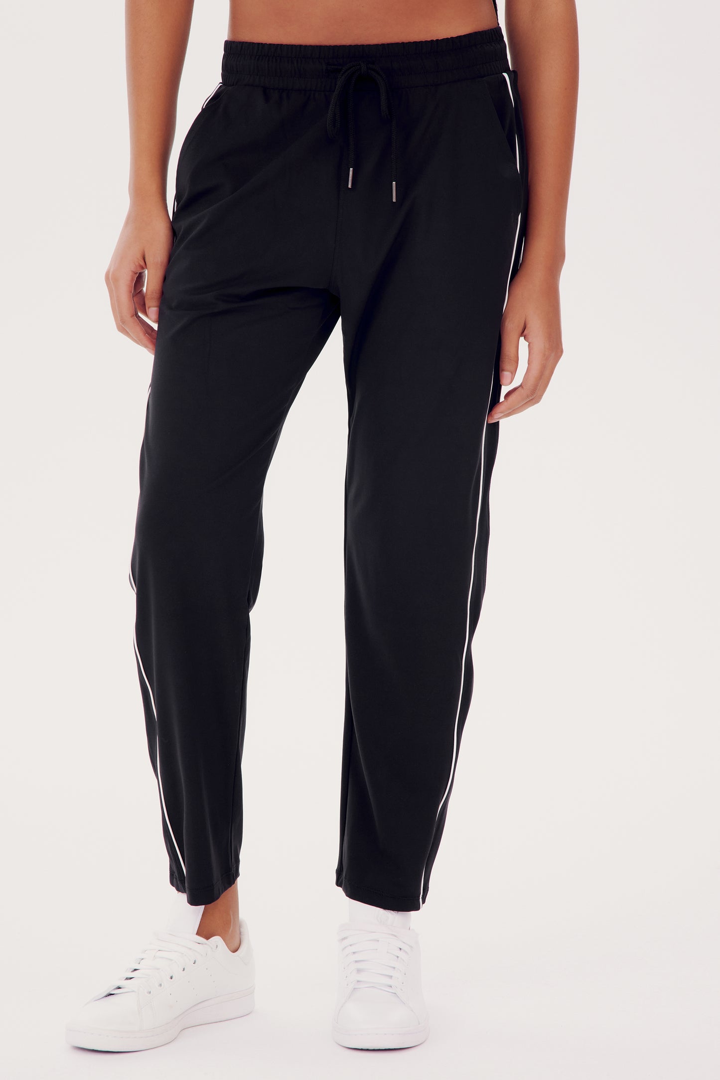 A person is wearing the Lucy Rigor Pant W/Piping - Black by SPLITS59, featuring relaxed fit and white side stripes, paired with white sneakers for all-day comfort.