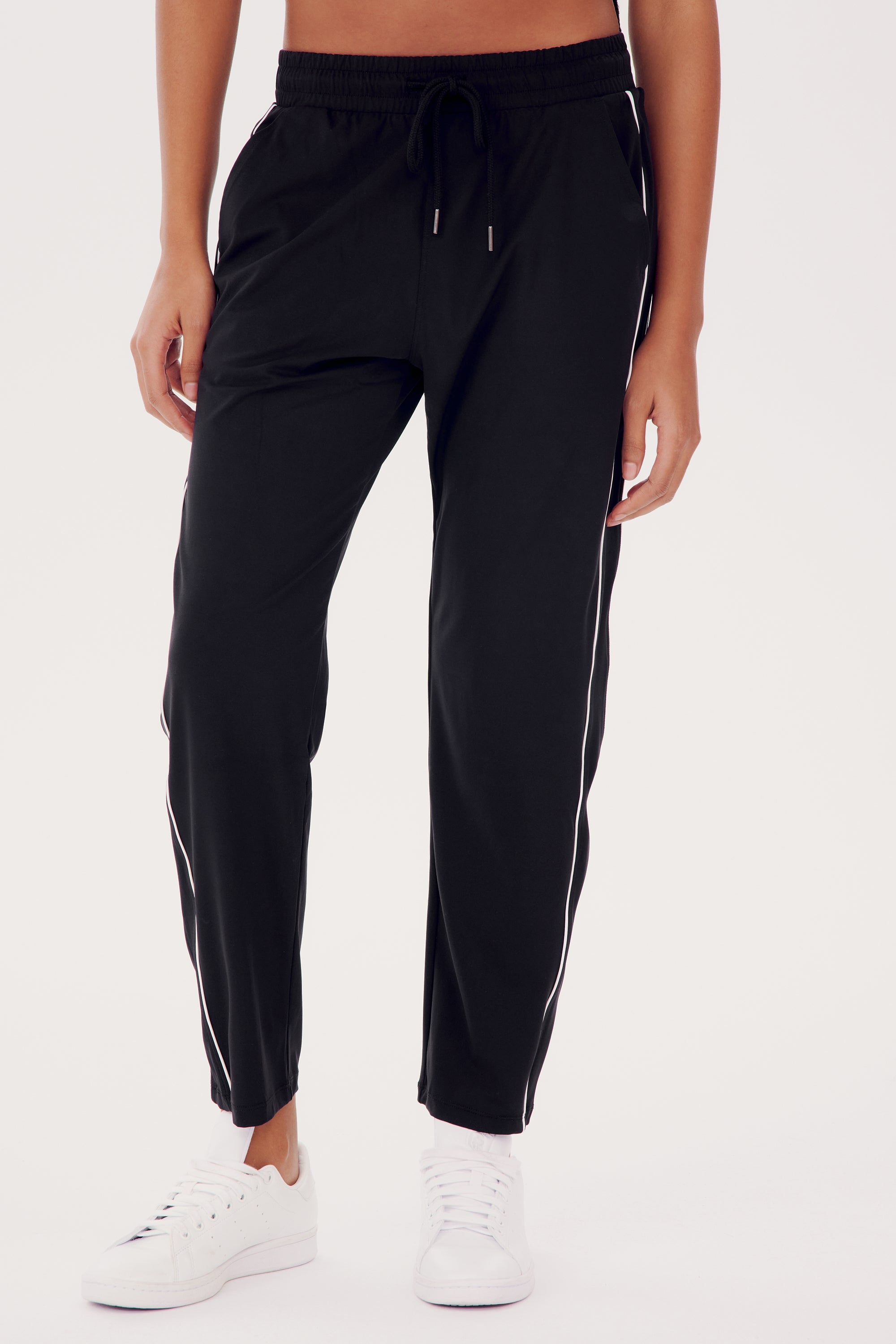 A person is wearing the Lucy Rigor Pant W/Piping - Black by SPLITS59, featuring relaxed fit and white side stripes, paired with white sneakers for all-day comfort.