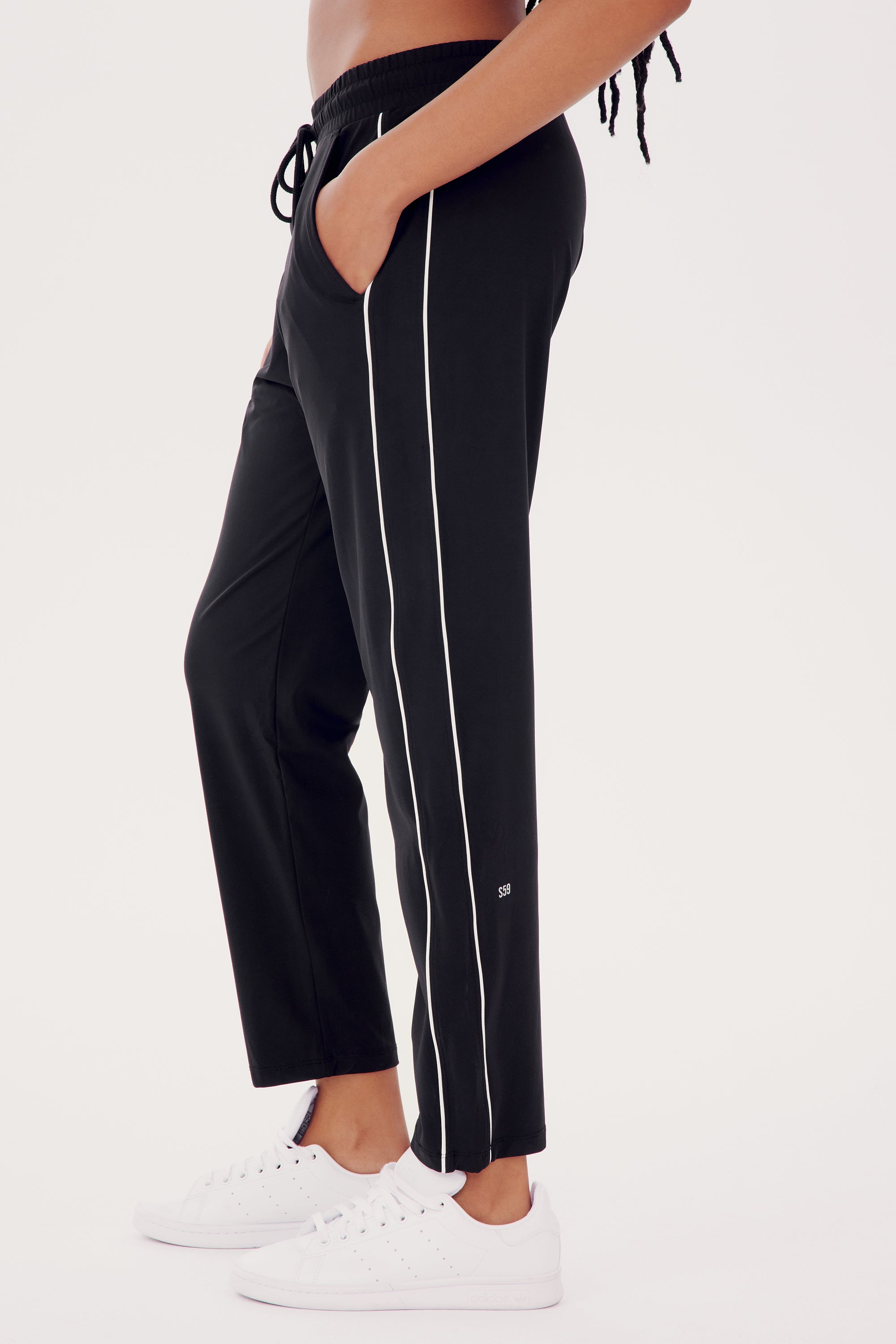 A person models the SPLITS59 Lucy Rigor Pant W/Piping in black, paired with white sneakers. With their hands in pockets and set against a plain backdrop, they showcase the jogger's all-day comfort and characteristic white stripe detail.