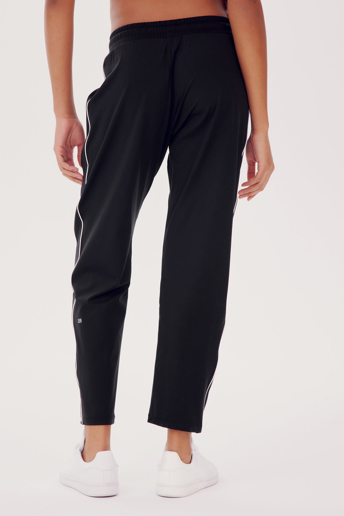 Wearing SPLITS59's Lucy Rigor Pant W/Piping in black, featuring white piping, a person stands with their back to the camera against a plain background. Paired with white sneakers, they embody all-day comfort.