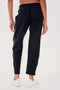 Wearing SPLITS59's Lucy Rigor Pant W/Piping in black, featuring white piping, a person stands with their back to the camera against a plain background. Paired with white sneakers, they embody all-day comfort.