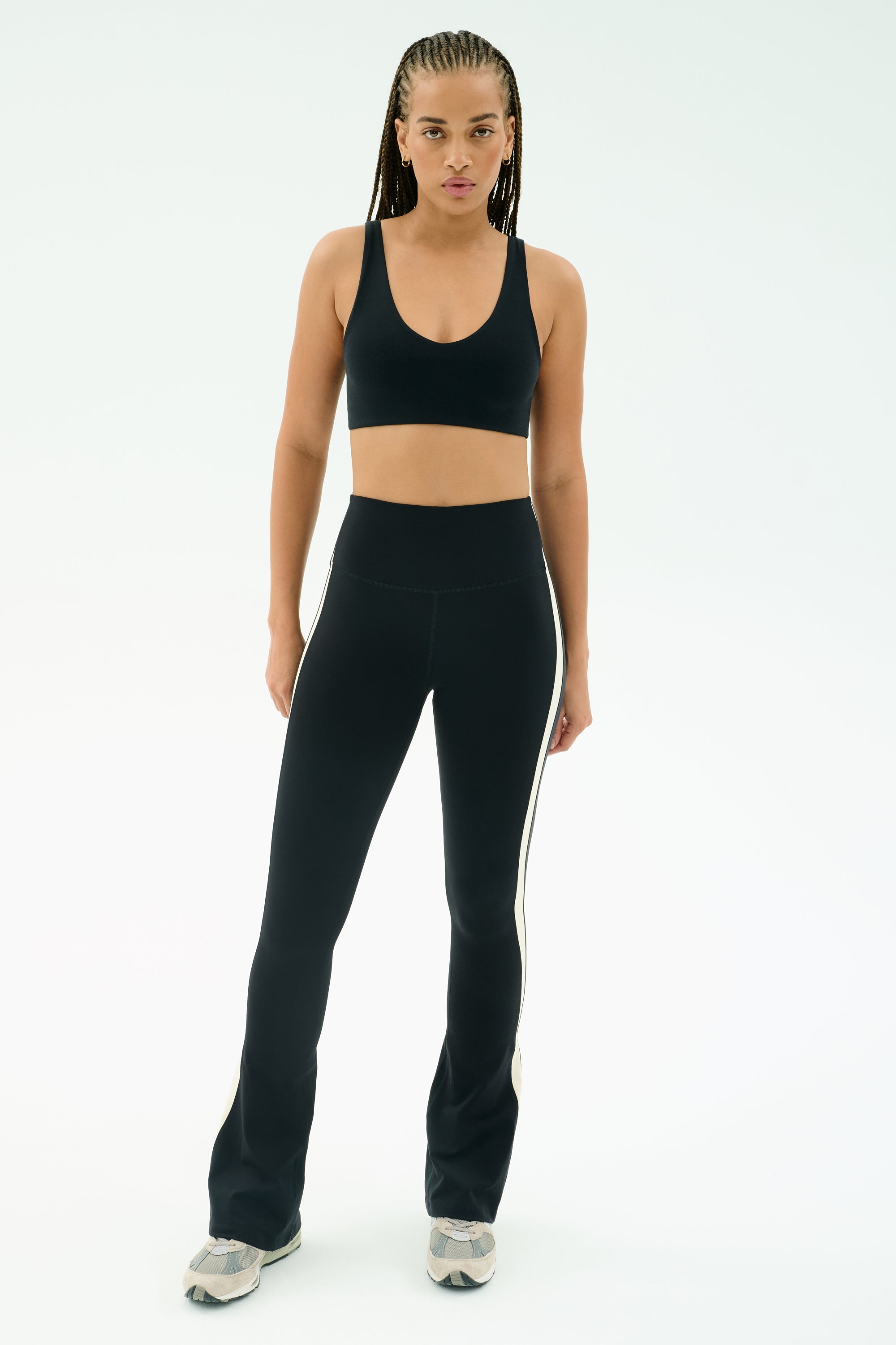 A person stands facing forward against a plain light background, wearing a black sports bra, SPLITS59 Raquel High Waist Flared Legging in Black/Creme with side stripes, and gray sneakers.
