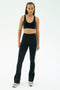A person stands facing forward against a plain light background, wearing a black sports bra, SPLITS59 Raquel High Waist Flared Legging in Black/Creme with side stripes, and gray sneakers.