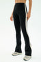 A person stands against a plain white background wearing SPLITS59 Raquel High Waist Flared Legging in Black/Creme, featuring a 4-way stretch, a white side stripe, and light-colored sneakers.
