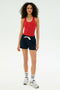 A woman faces forward against a plain background, wearing the SPLITS59 Reena Sweatshort in Indigo, a red tank top, white socks with black stripes, and white sneakers.