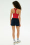 A person stands facing away, wearing a red racerback top, SPLITS59 Reena Sweatshort in Indigo, white socks, and sneakers against a plain light background.