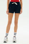 A person wears a red top, SPLITS59 Reena Sweatshort - Indigo, white striped socks, and white sneakers, standing against a plain background.