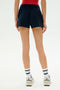 A person stands facing away, wearing a red top, white crew socks with blue stripes, white sneakers, and SPLITS59’s Reena Sweatshort in Indigo against a plain background.