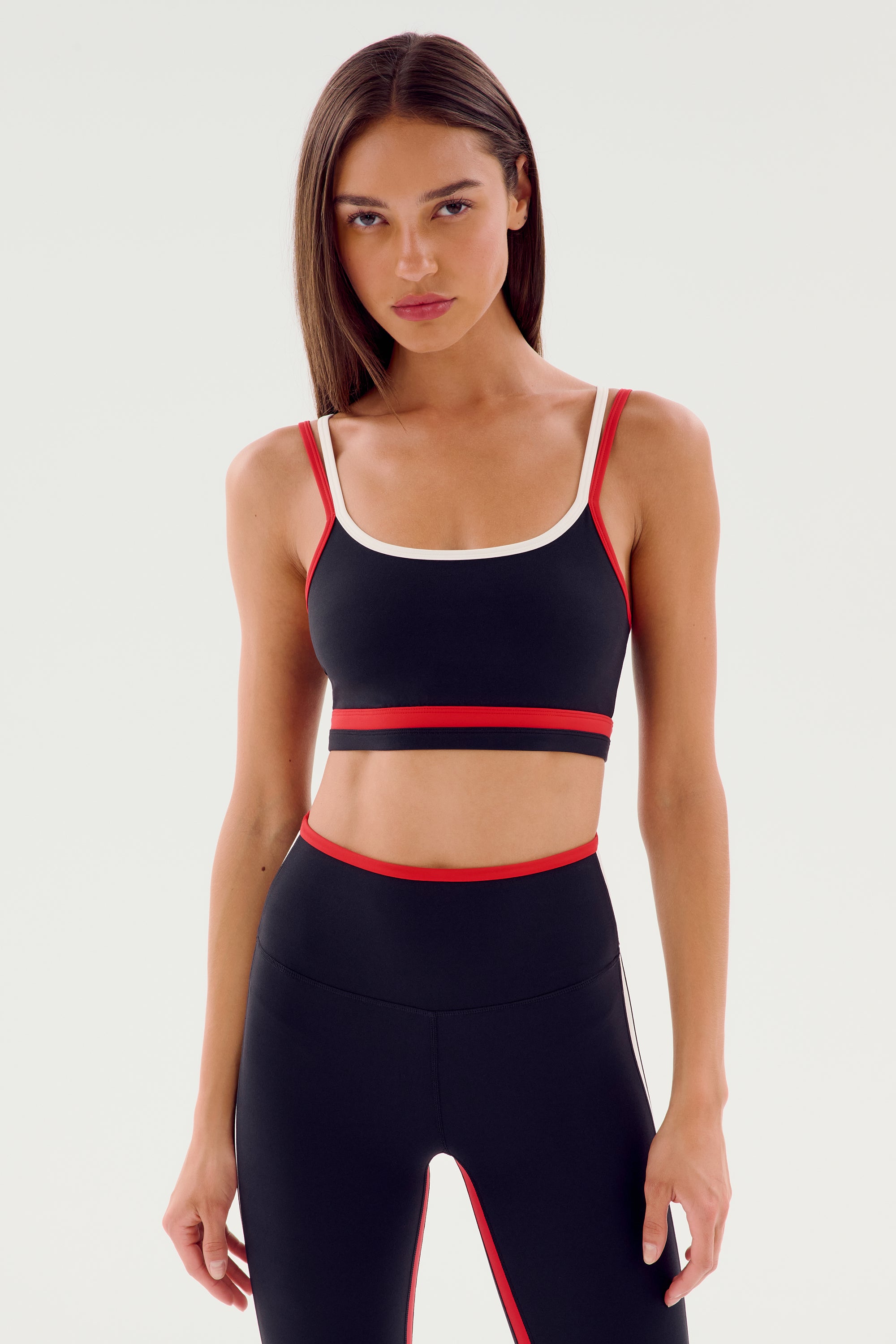 A woman is wearing a black SPLITS59 Eva Rigor Double Strap Bra - Black/Pirate Red and leggings with red and white trim, standing comfortably confident against a plain white background.