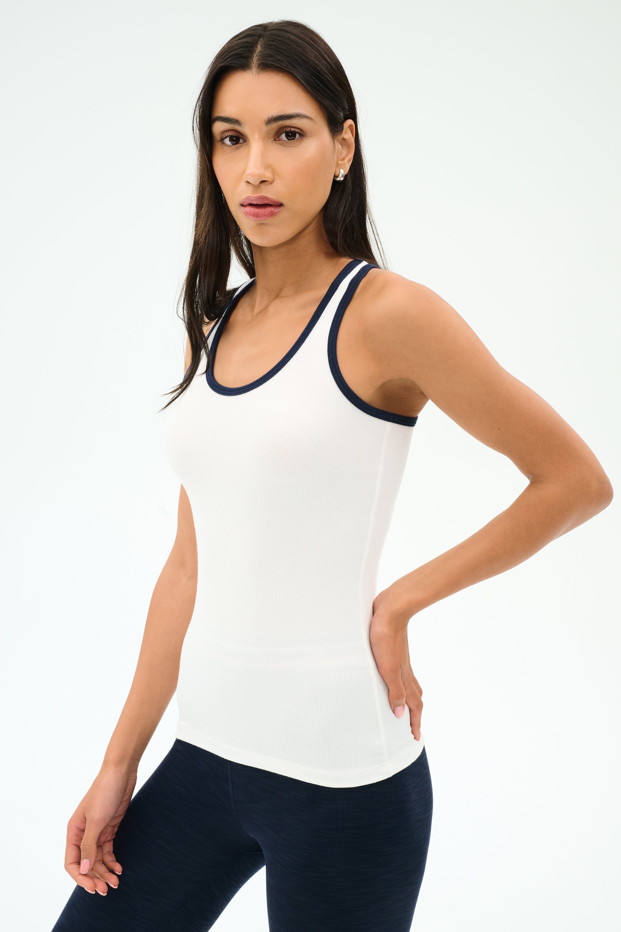 A woman stands against a plain background, wearing the SPLITS59 Ashby Rib Tank in White/Indigo and navy leggings, looking at the camera with a neutral expression—ideal for yoga or gym workouts.