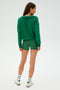A person with long hair is turned away, wearing an SPLITS59 Joan Fleece Polo Sweatshirt in Arugula, shorts, white sneakers, and striped socks, standing against a plain background.