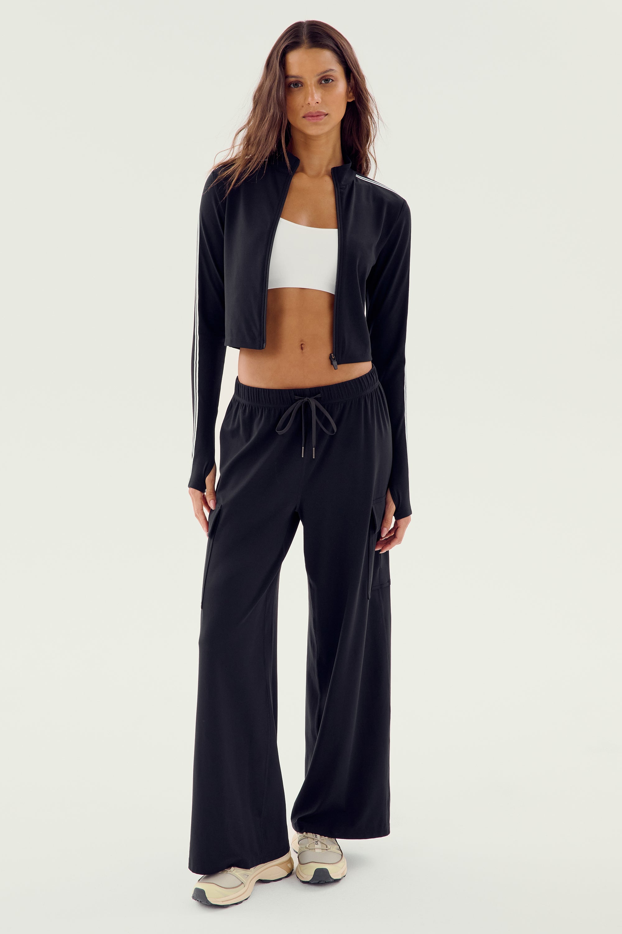 A woman stands facing forward, wearing a black Leo Airweight Jacket Crop from SPLITS59, black drawstring pants made of 81% nylon and 19% spandex, a white sports bra, and sneakers.