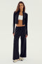A woman stands facing forward, wearing a black Leo Airweight Jacket Crop from SPLITS59, black drawstring pants made of 81% nylon and 19% spandex, a white sports bra, and sneakers.