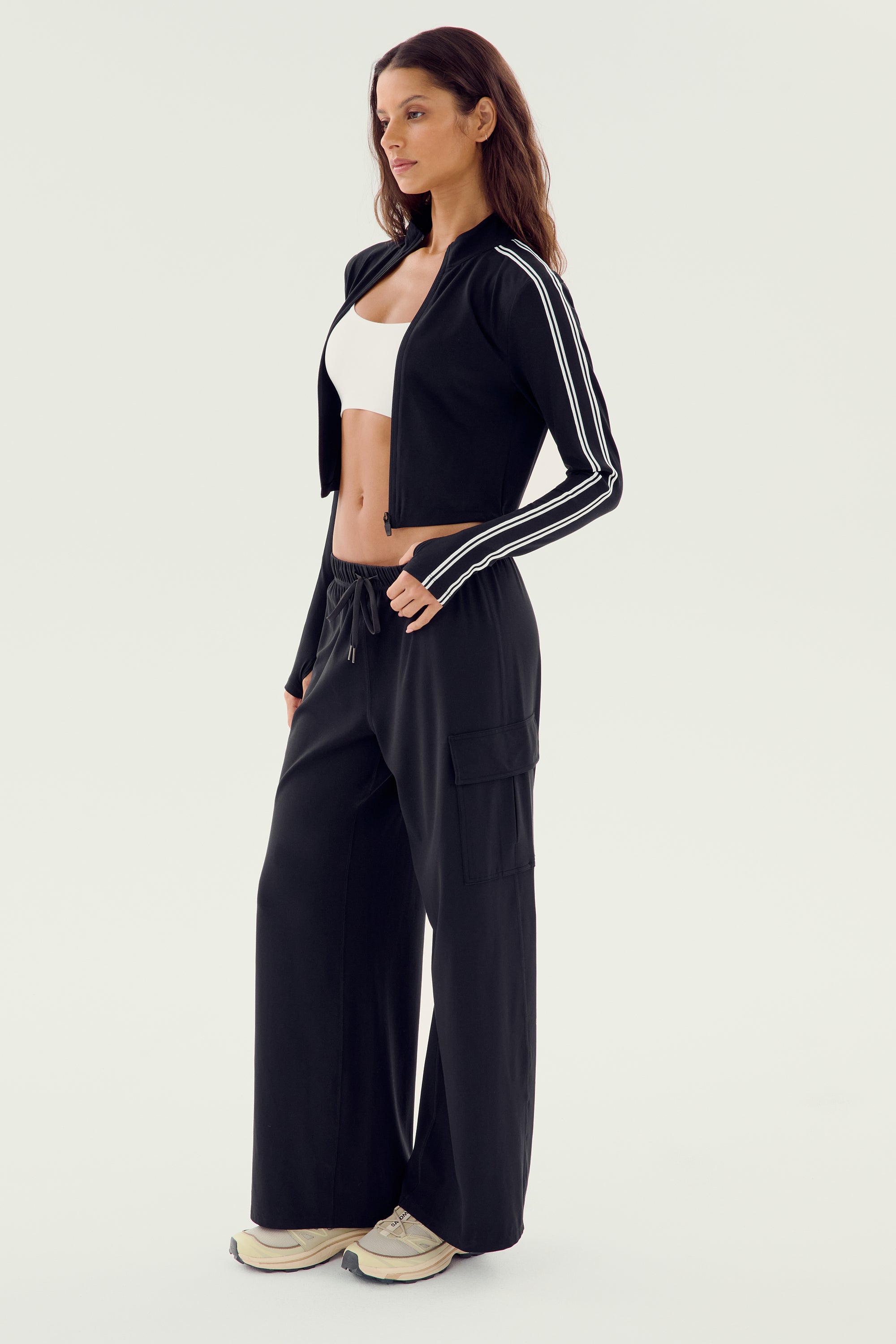 A woman in a white sports bra and the SPLITS59 Leo Airweight Jacket Crop - Black/White stands in profile. She wears black wide-leg pants with pockets and white sneakers. Made of 19% spandex and 81% nylon, this outfit offers both style and comfort.