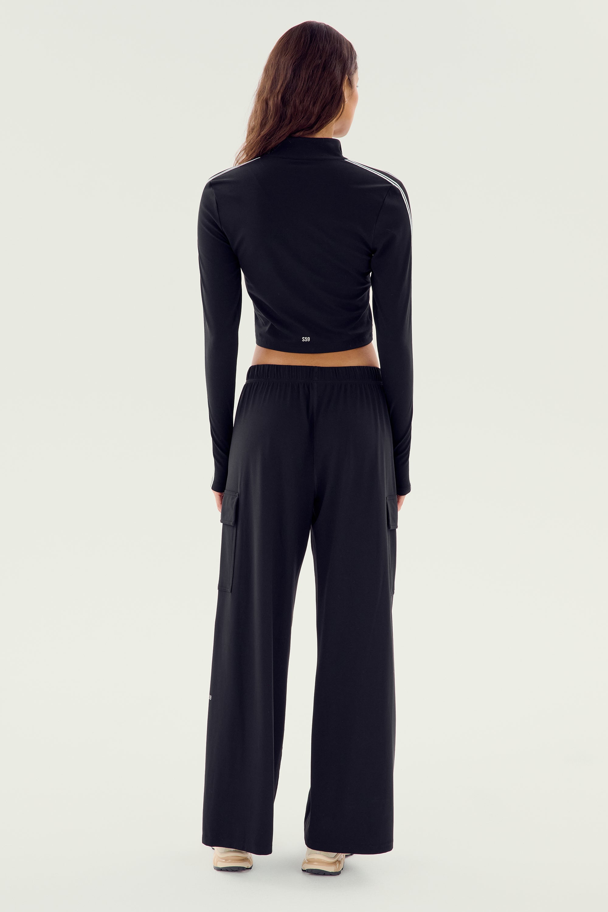 A person stands facing away, wearing the SPLITS59 Leo Airweight Jacket Crop in a sleek black and white colorway, paired with wide-leg pants. Both garments are made from a blend of 19% spandex and 81% nylon. They have long hair and are set against a plain background.
