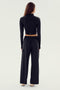 A person stands facing away, wearing the SPLITS59 Leo Airweight Jacket Crop in a sleek black and white colorway, paired with wide-leg pants. Both garments are made from a blend of 19% spandex and 81% nylon. They have long hair and are set against a plain background.