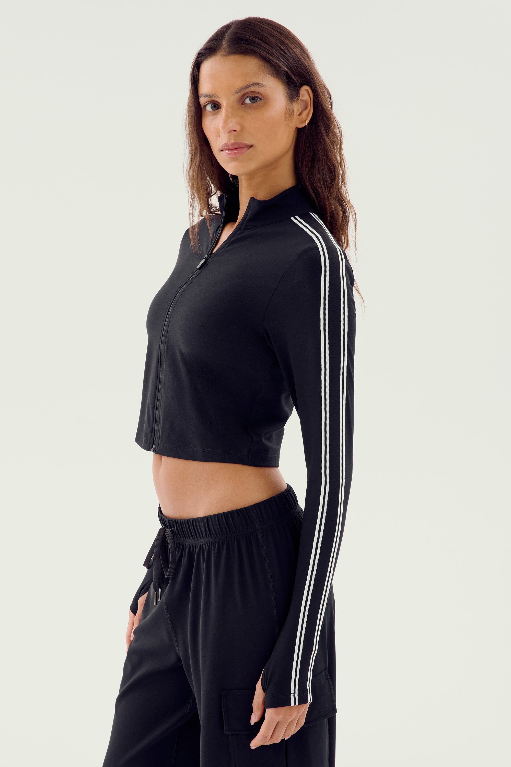 A person with long hair stands in profile wearing the Leo Airweight Jacket Crop in black and white by SPLITS59, paired with matching black nylon pants.
