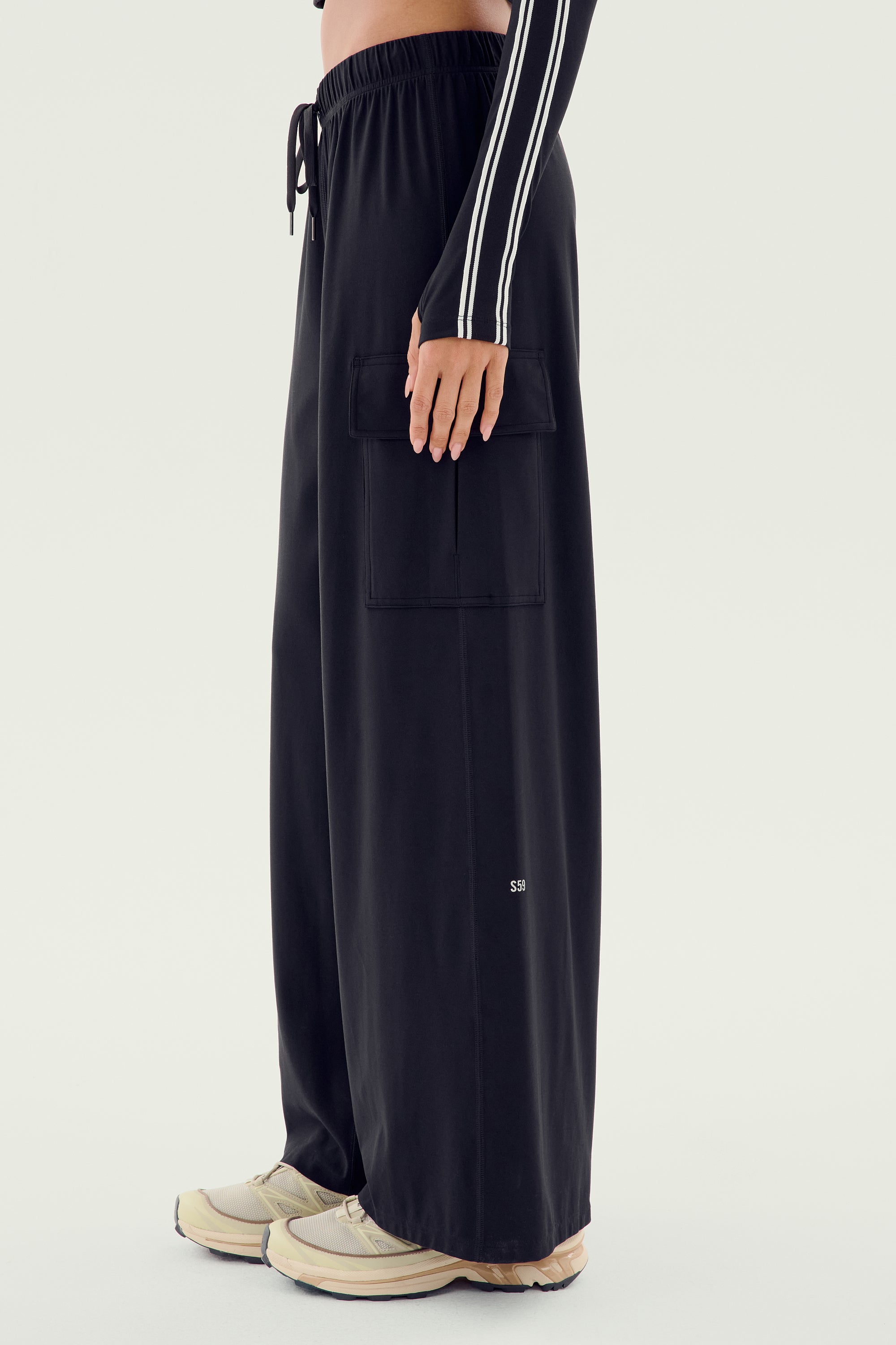 A person wearing the SPLITS59 Alex Airweight Cargo Pant in Black, which features a pocket on the side, a long-sleeve top with white stripes on the arms, and white sneakers.