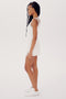 A woman with long braided hair stands in profile wearing a white sleeveless SPLITS59 Martina Rigor Dress w/Piping - White and white sneakers against a plain white background.