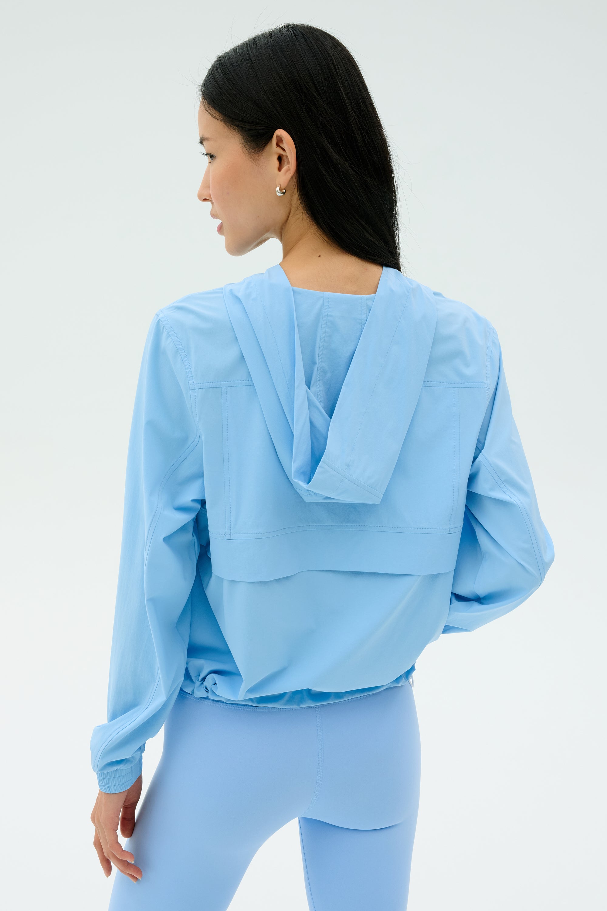 A woman with long black hair is seen from behind wearing the SPLITS59 Megan Woven Nylon Jacket in Skylight, paired with matching leggings, both crafted from soft nylon-spandex fabric against a plain background.