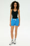 A person stands facing forward wearing SPLITS59's Reena Sweatshort in Riviera, a black tank top, striped socks, and white sneakers against a plain background.