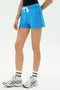 Person wears SPLITS59's Reena Sweatshort - Riviera in bright blue, paired with white socks and sneakers, standing against a plain light background.
