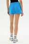 A person wearing SPLITS59 Reena Sweatshort - Riviera, white socks, and running shoes stands with their back to the camera against a plain background.