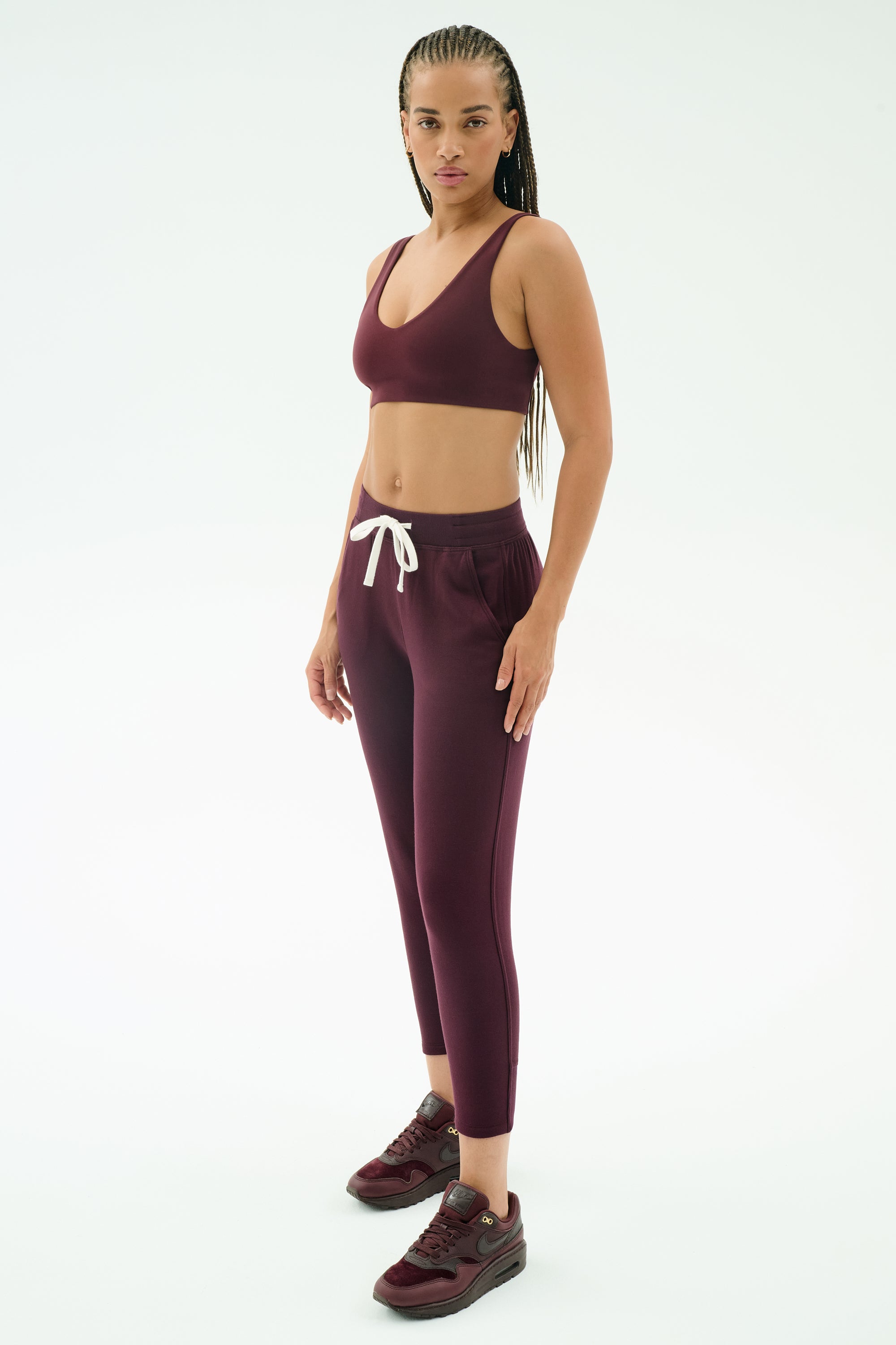 A woman stands against a white background wearing SPLITS59 Reena Fleece 7/8 Sweatpant in Dark Cherry, paired with a matching maroon sports bra and sneakers.