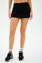 A person wearing the Sally Rigor Skort from SPLITS59, paired with black and white sneakers and white socks featuring black stripes, stands against a plain background.