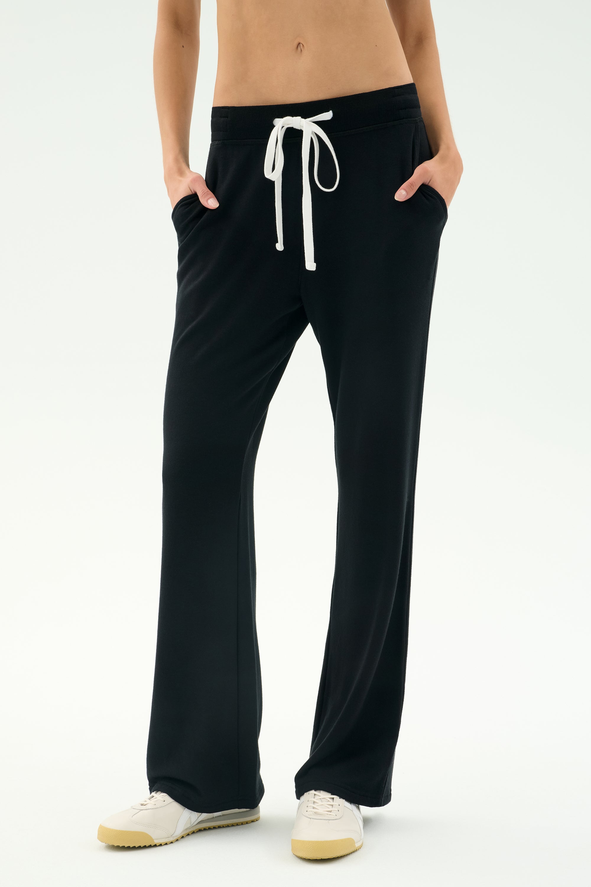 A person in SPLITS59 Raven Fleece Full Length Sweatpant - Black and white sneakers stands with hands in pockets against a plain background.