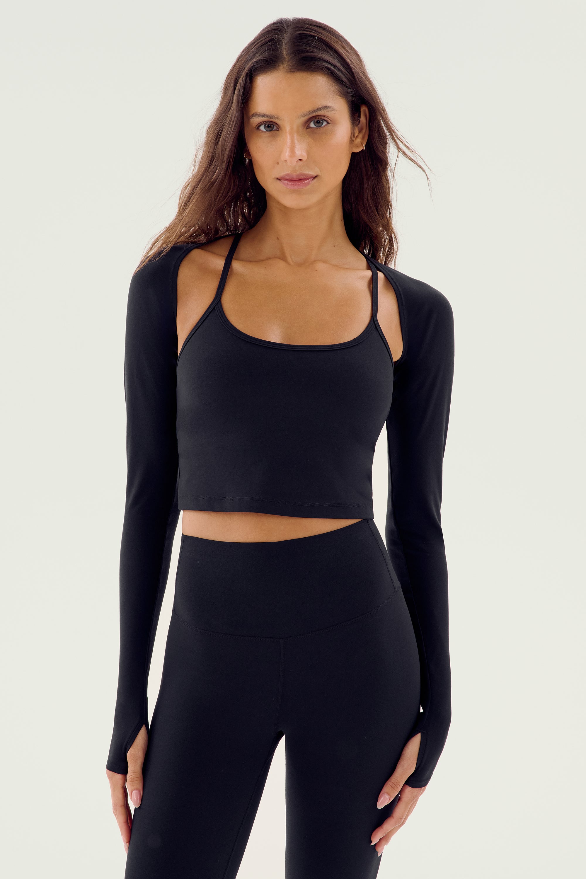 A woman with long brown hair is wearing the SPLITS59 Shiri Airweight Shrug - Black, a stylish long-sleeve crop top with thumbholes, paired with black high-waisted leggings. Both pieces are crafted from a comfortable blend of nylon and spandex. She stands facing forward against a plain white background.