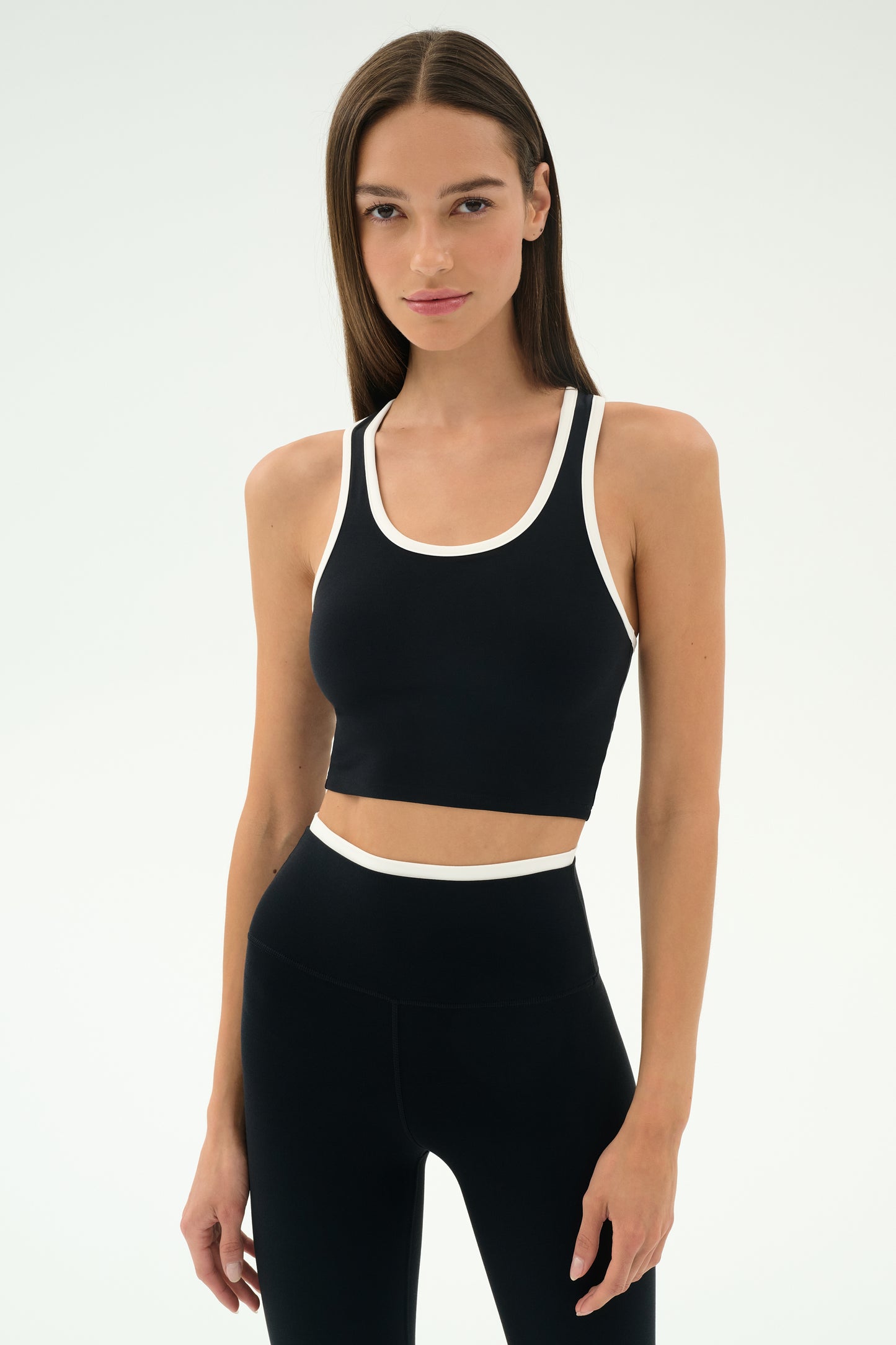 A woman with straight brown hair wears the SPLITS59 Amber Airweight Bralette in Black/White, a cropped scoop-neck athletic top made from Airweight performance fabric, perfect for hot yoga, against a plain light background.