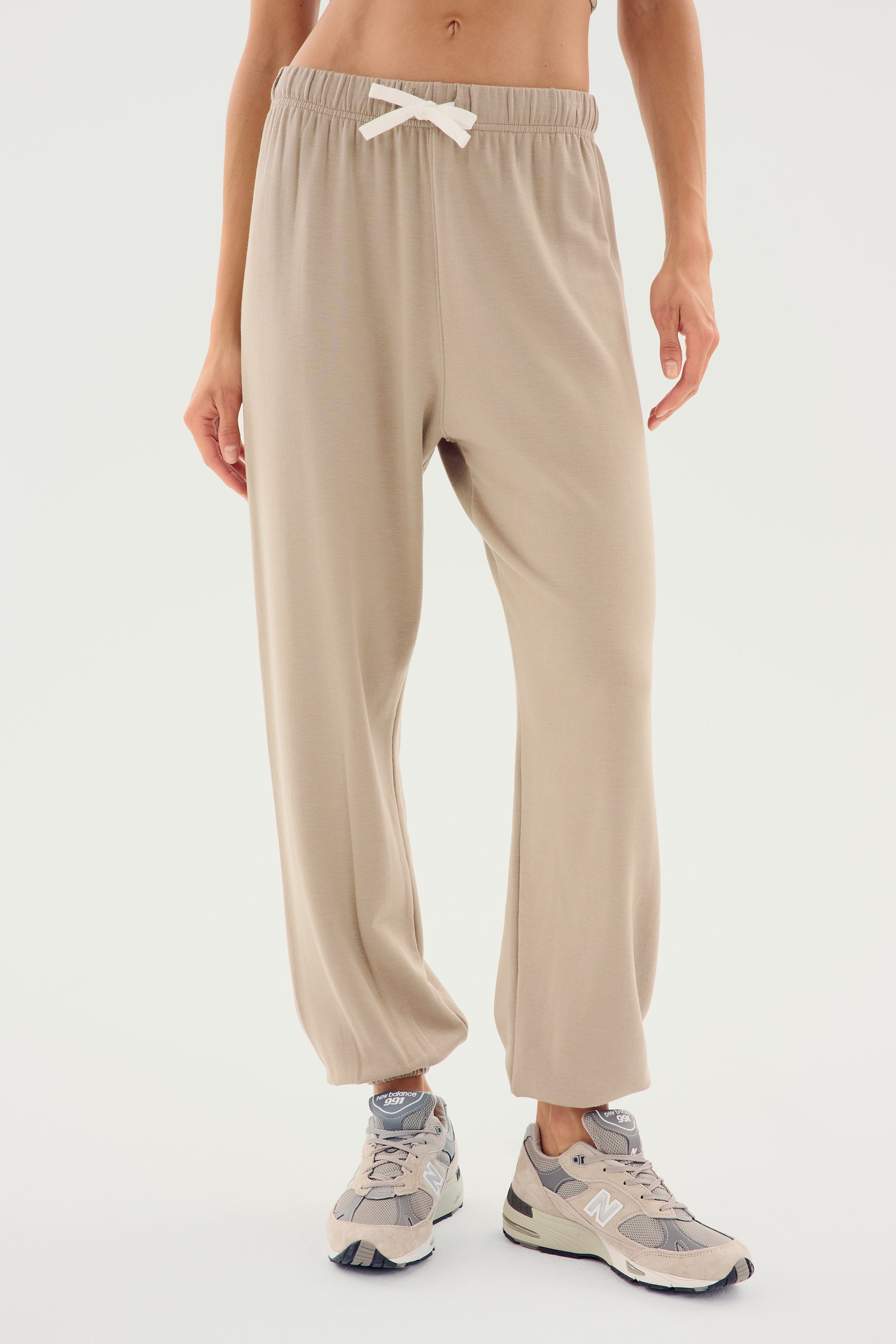 A person is shown from the chest down, wearing SPLITS59 Andie Oversized Fleece Sweatpant - Latte with a white drawstring. They are also wearing gray sneakers with white laces, perfect for cool weather workouts. The background is plain white.