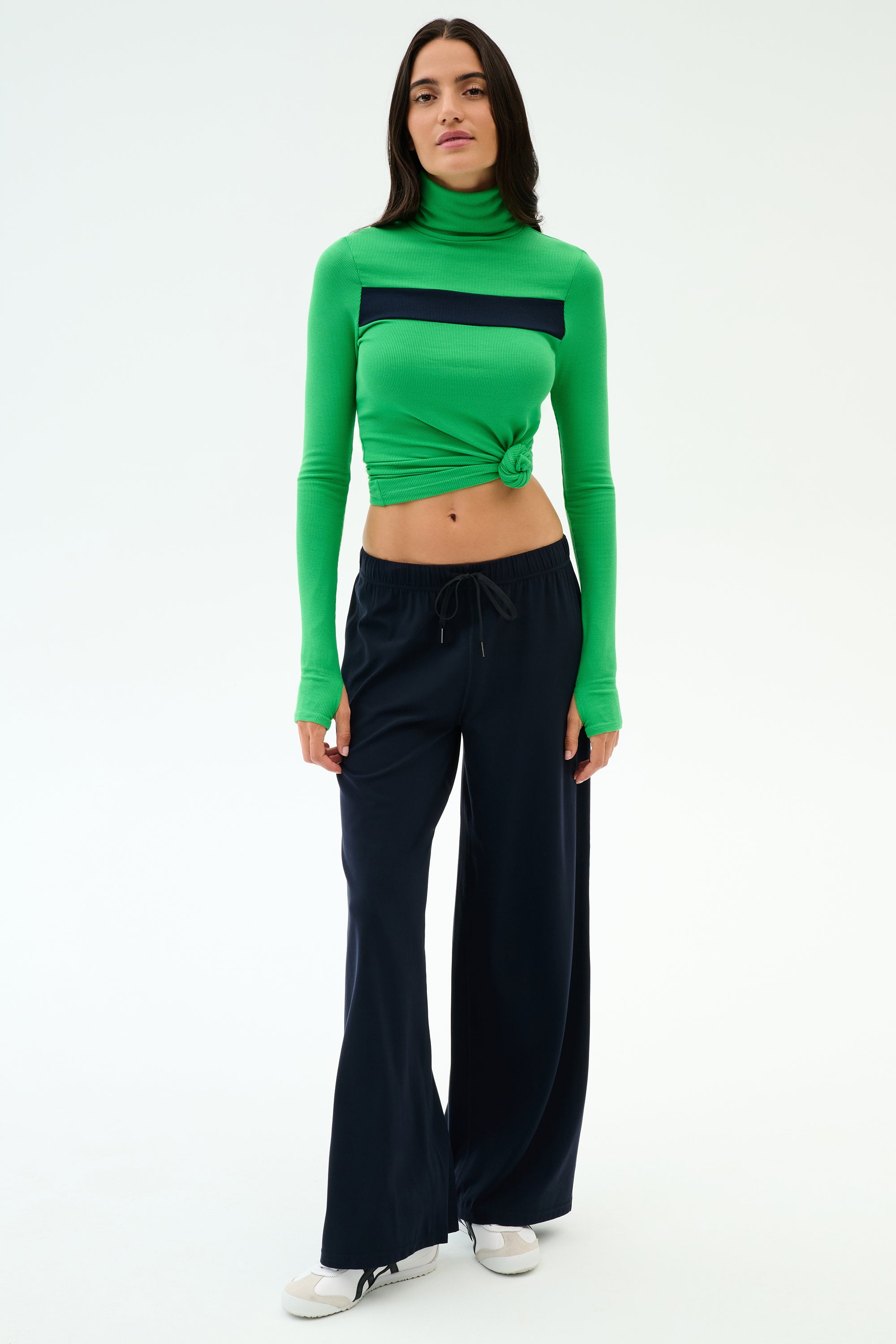 Woman stands against a plain background, wearing a green long-sleeve turtleneck with front knot, SPLITS59 Brynn Airweight Pant in Indigo, and white sneakers.