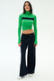 Woman stands against a plain background, wearing a green long-sleeve turtleneck with front knot, SPLITS59 Brynn Airweight Pant in Indigo, and white sneakers.