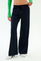 A person wears SPLITS59 Brynn Airweight Pant in Indigo, featuring a comfortable nylon-spandex blend, paired with a green long-sleeve top and white sneakers, standing against a plain white background.
