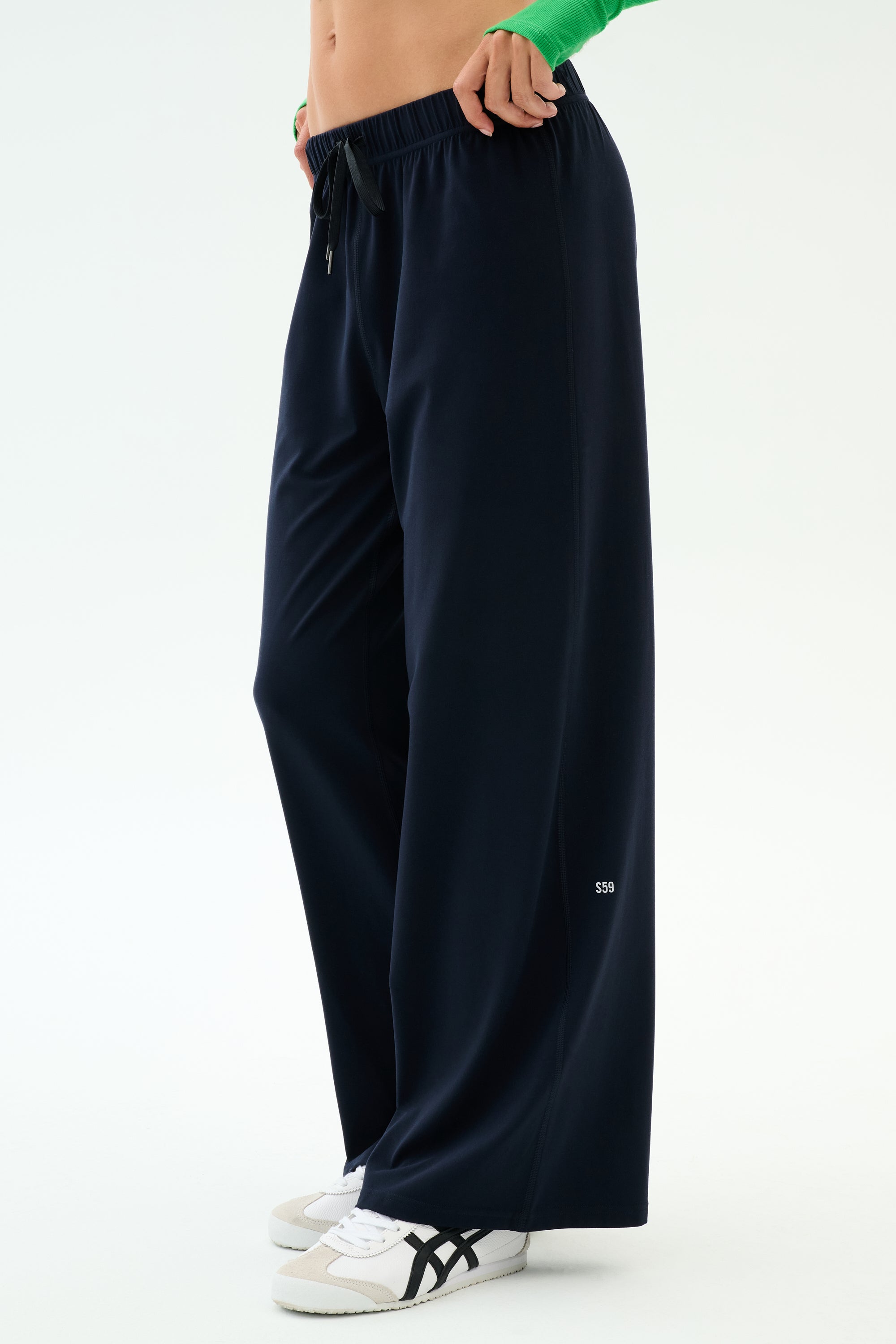 Person wearing SPLITS59 Brynn Airweight Pant in Indigo, paired with white sneakers featuring black stripes and a green long-sleeve top, standing against a plain light background.