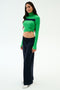 A woman wears the SPLITS59 Brynn Airweight Pant in Indigo with a green long-sleeve turtleneck top, tied at the waist, and white sneakers, standing against a plain white background.
