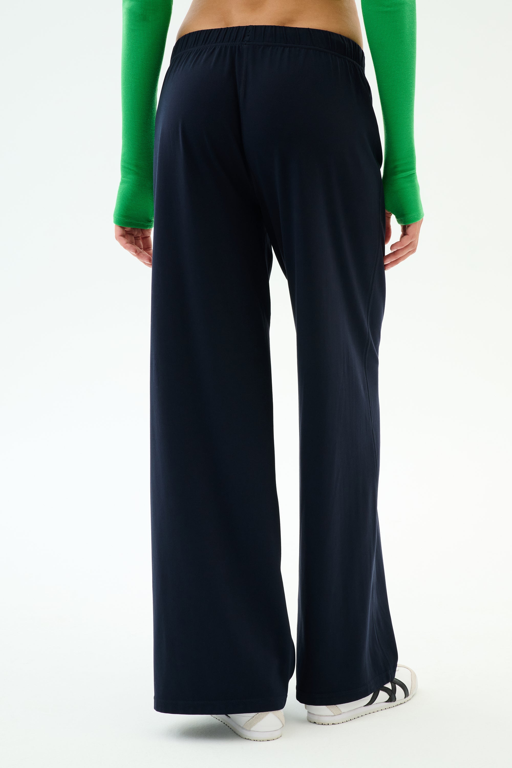 A person stands with their back to the camera against a plain background, wearing SPLITS59's Brynn Airweight Pant in Indigo, paired with a green long-sleeve top and white sneakers.