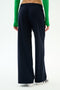 A person stands with their back to the camera against a plain background, wearing SPLITS59's Brynn Airweight Pant in Indigo, paired with a green long-sleeve top and white sneakers.