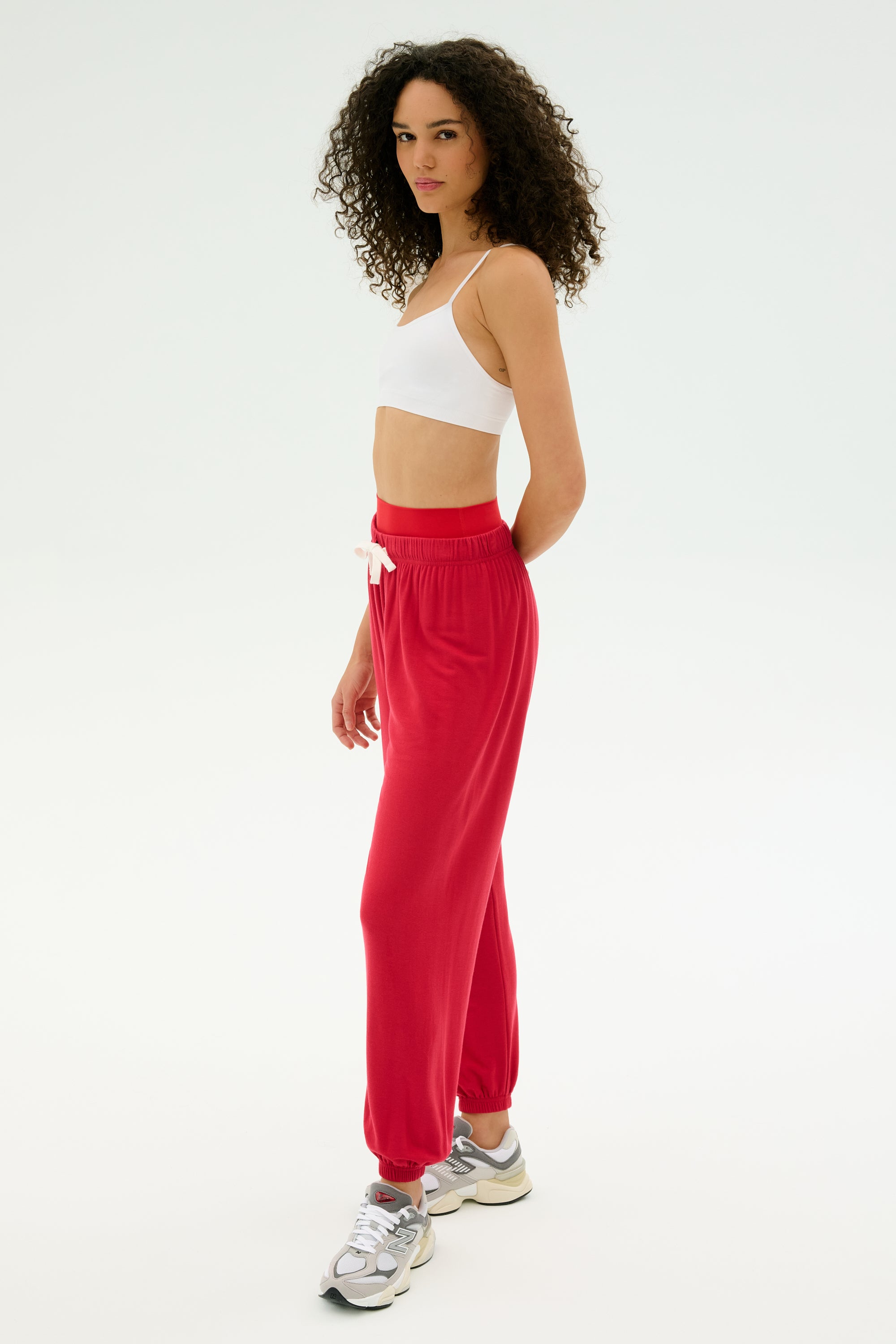 A person with curly hair wears a white crop top, SPLITS59’s Andie Oversized Fleece Sweatpant in Pirate Red, and gray sneakers in a minimalist white studio—chic cool-weather inspiration for workouts or casual warm-ups.